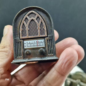 May include: A miniature antique-style radio with a dark brown finish. The radio features a decorative arched top with a mesh grille and a dial with numbers. Two knobs are visible below the dial. The radio's dimensions are not visible.