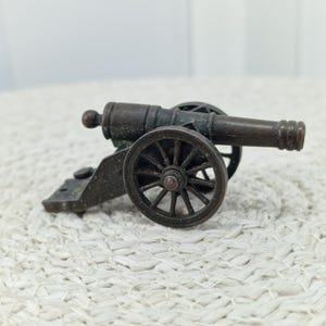 May include: A miniature bronze cannon with spoked wheels and a detailed barrel. The small artillery piece is a decorative item, likely a collectible or a model. The cannon is set against a neutral background.