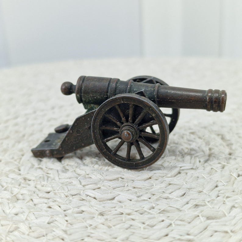 May include: A miniature bronze cannon with spoked wheels and a detailed barrel. The small artillery piece is a decorative item, likely a collectible or a model. The cannon is set against a neutral background.