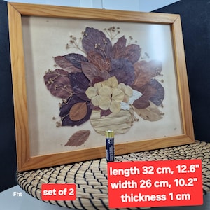 May include: A framed botanical art piece featuring a dried flower arrangement. The artwork includes a variety of dried leaves and flowers in shades of brown, purple, and cream. The frame is made of light-colored wood. Dimensions: 12.6" length, 10.2" width, and 0.4" thickness.