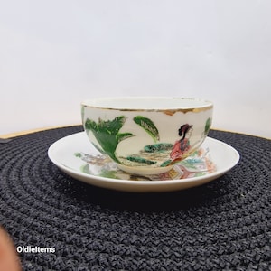 Peut inclure: Une tasse à thé et une soucoupe en porcelaine blanche avec un motif floral vert, rose et rouge représentant une femme en robe traditionnelle chinoise. La tasse et la soucoupe ont un bord doré.