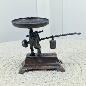 Pode incluir: Uma pequena balança de bronze em estilo vintage com uma bandeja redonda, um braço de equilíbrio e um peso suspenso. A balança é montada numa base retangular com pés decorativos. O design sugere que é um item em miniatura ou decorativo.