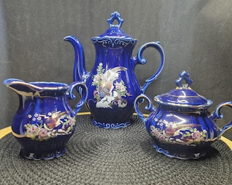 May include: A cobalt blue porcelain tea set featuring a teapot, creamer, and sugar bowl. Each piece is adorned with a gold trim and a detailed floral and bird design. The set is displayed on a black surface.