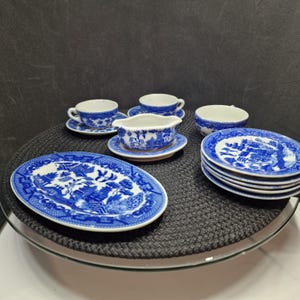Può includere: Una collezione di piatti in porcellana blu e bianca, tra cui tazze, piattini, piatti e una salsiera. I piatti presentano un classico motivo Willow. Il set è disposto su una superficie nera.