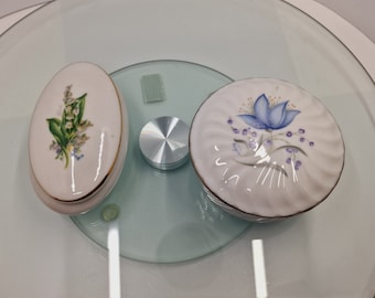 May include: Two small porcelain trinket boxes with floral designs. One box features a white background with a blue flower and purple accents. The other box features a white background with a green and white lily of the valley design.