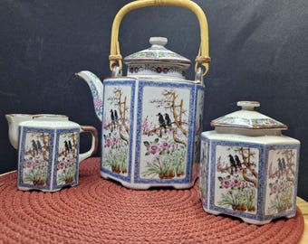 May include: A white porcelain tea set with a blue and floral design. The set includes a teapot with a bamboo handle, a creamer, and a sugar bowl. The teapot has a lid with a knob in the shape of a bird. The creamer and sugar bowl have lids with knobs in the shape of birds. The set is decorated with a floral design and birds.
