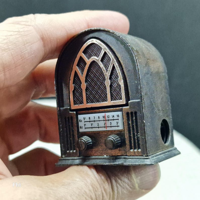 May include: A miniature antique-style radio in shades of brown and bronze. The radio features a detailed facade with a speaker grill, a tuning dial, and a frequency scale. The radio is held in a hand.