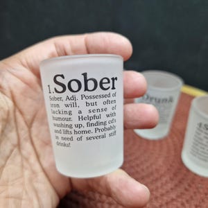 May include: A frosted glass shot glass with the word "Sober" printed in black, along with a definition. The shot glass is held in a hand, with two other shot glasses visible in the background.