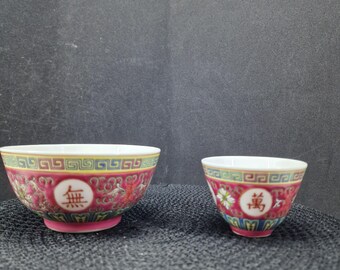 May include: Two porcelain bowls with a red and gold floral design. The larger bowl has the Chinese character "無" and the smaller bowl has the character "萬". The bowls have a pink base and a white interior.