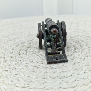 May include: A miniature bronze cannon with two wheels and a detailed carriage. The cannon barrel is cylindrical, and the wheels are circular. The overall design suggests a historical or antique theme.