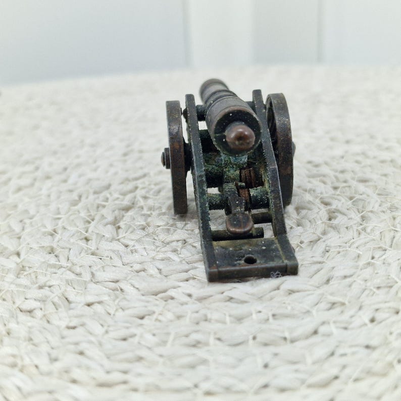 May include: A miniature bronze cannon with two wheels and a detailed carriage. The cannon barrel is cylindrical, and the wheels are circular. The overall design suggests a historical or antique theme.