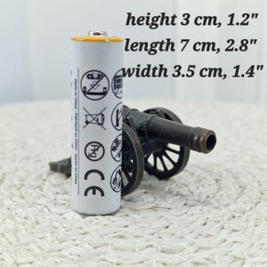 May include: A white battery with a yellow top stands next to a miniature bronze cannon. The battery is 1.2 inches high, 2.8 inches long, and 1.4 inches wide. The cannon has two wheels and a detailed barrel. The battery has several symbols printed on it.