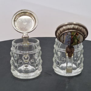 May include: Two miniature glass beer steins with silver lids. One lid has a small image of a building and trees. Both steins have a textured, bubble-like design.
