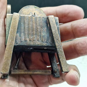 May include: A miniature antique camera model with a bronze finish. The camera features a detailed design with a lens, viewfinder, and tripod-like legs. The model is held in a hand, showcasing its small size and intricate craftsmanship. The text "Fht" is visible.