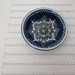 K&ouml;nnte beinhalten: Eine kleine, blaue Keramiksch&uuml;ssel mit wei&szlig;em Rand und handbemaltem Blumendesign. Das Design zeigt rosa, gelbe und gr&uuml;ne Blumen mit wei&szlig;en Umrissen.