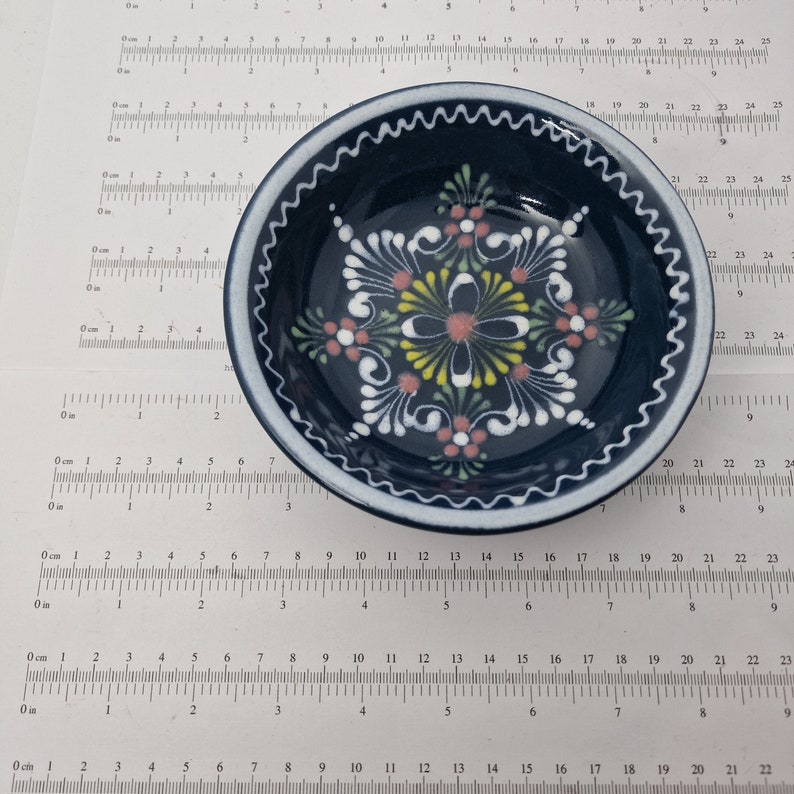 K&ouml;nnte beinhalten: Eine kleine, blaue Keramiksch&uuml;ssel mit wei&szlig;em Rand und handbemaltem Blumendesign. Das Design zeigt rosa, gelbe und gr&uuml;ne Blumen mit wei&szlig;en Umrissen.