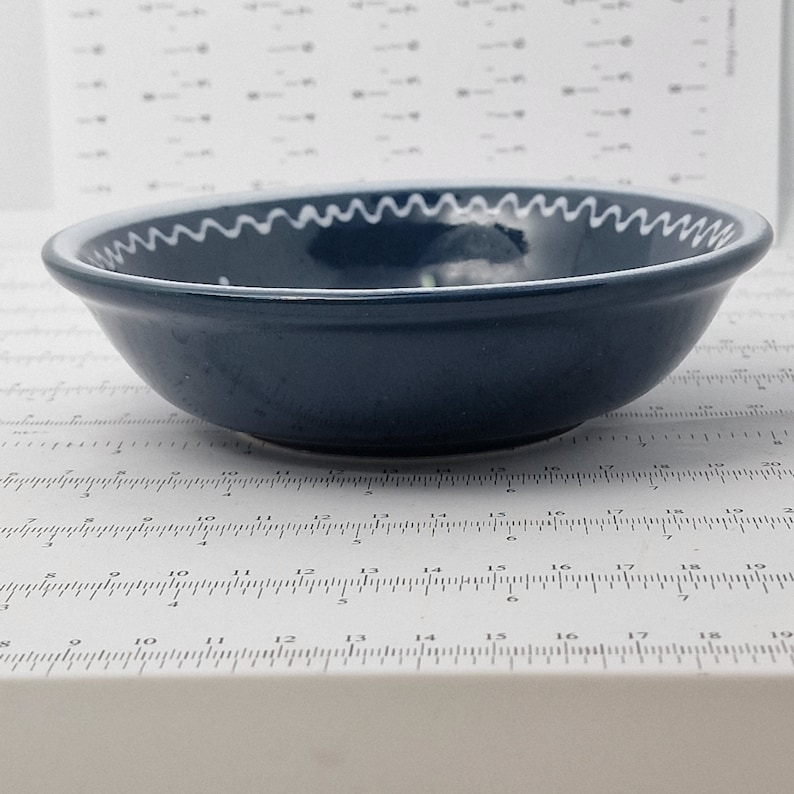 K&ouml;nnte beinhalten: Eine dunkelblaue Keramiksch&uuml;ssel mit einem wei&szlig;en, gewellten Rand. Die Sch&uuml;ssel ist leer und steht auf einer wei&szlig;en Oberfl&auml;che.