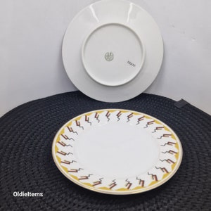 May include: Two white ceramic plates with a gold rim. The top plate has a geometric design in brown and yellow. The bottom plate has a green mark with the text "Franz" in black.