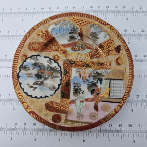 Può includere: Un piccolo piattino rotondo in ceramica con un bordo dorato e intricati disegni di ispirazione giapponese. Il piattino presenta una scena centrale con due figure in abiti tradizionali, circondate da motivi floreali e geometrici.