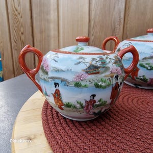 Puede incluir: Un azucarero de porcelana blanca con tapa y asas rojas. El bol está decorado con una escena de inspiración japonesa con un puente, montañas y flores de cerezo. También hay figuras de mujeres con ropa tradicional japonesa.