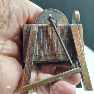 May include: A miniature bronze-colored camera, featuring a detailed design with vertical lines and a circular top. The camera has a small lever and a rectangular base. The letters "Fht" are visible on the bottom left.