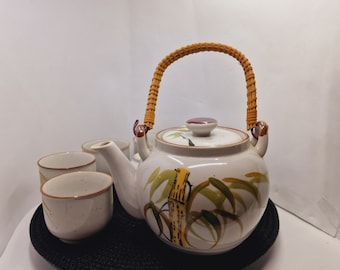 May include: A white ceramic teapot with a bamboo design and a brown wicker handle. The teapot is on a black round tray with four white ceramic teacups.
