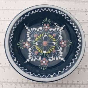 K&ouml;nnte beinhalten: Eine blaue Keramiksch&uuml;ssel mit einem wei&szlig;en gewellten Rand und einem handgemalten Blumendesign in Wei&szlig;, Gelb, Rosa und Gr&uuml;n.
