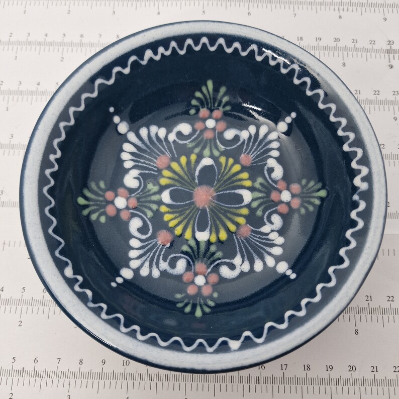 K&ouml;nnte beinhalten: Eine blaue Keramiksch&uuml;ssel mit einem wei&szlig;en gewellten Rand und einem handgemalten Blumendesign in Wei&szlig;, Gelb, Rosa und Gr&uuml;n.