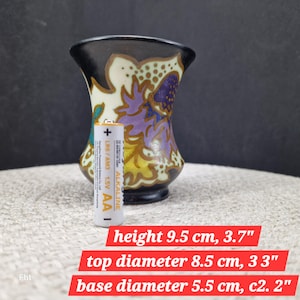 May include: A small, decorative vase with a black rim and base, featuring a floral design in purple, yellow, and white. The vase is 3.7 inches tall, with a top diameter of 3.3 inches and a base diameter of 2.2 inches. An AA battery is next to the vase.