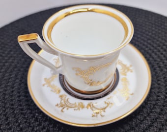 May include: A white porcelain teacup and saucer with gold trim and a gold floral design. The cup has a single handle.