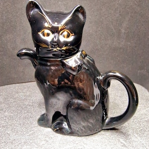 May include: Black ceramic teapot shaped like a cat with gold accents. The cat has a bow tie and a raised tail.