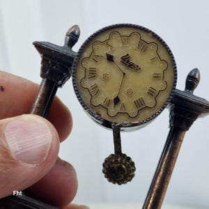 Op de afbeelding: Een miniatuur antieke klok met een crèmekleurige wijzerplaat en Romeinse cijfers. Op de wijzerplaat staat het woord "Urgos". De klok wordt vastgehouden door twee sierlijke, bronskleurige steunen en heeft een decoratieve slinger.