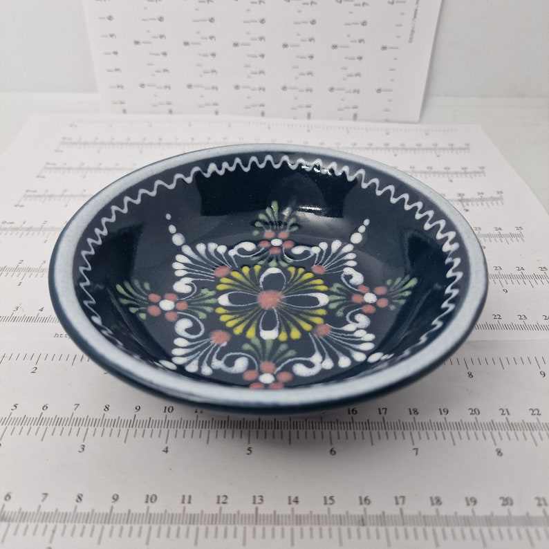 K&ouml;nnte beinhalten: Eine kleine, blaue Keramiksch&uuml;ssel mit wei&szlig;em Rand und einem wei&szlig;en Blumenmuster in der Mitte. Das Muster hat rosa, gelbe und gr&uuml;ne Akzente.