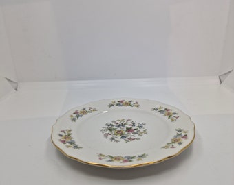 May include: A white ceramic plate with a scalloped edge and a floral design in shades of pink, yellow, and blue. The plate has a gold trim around the edge.