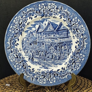 Puede incluir: Plato decorativo azul y blanco con un borde floral y una escena detallada de una plaza. El plato presenta una imagen central de edificios y personas, con una estética clásica y vintage. El plato se exhibe sobre un soporte dorado.