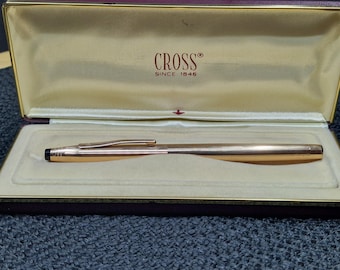 May include: A gold-colored Cross pen in an open burgundy and gold presentation box. The pen is displayed on a cream-colored interior. The box lid features the Cross logo and the text "SINCE 1846".