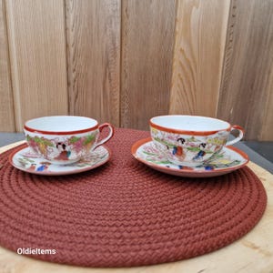 Op de afbeelding: Twee vintage theekopjes en schoteltjes met een Japans geisha-ontwerp. De kopjes en schoteltjes zijn wit met een rode rand en hebben een kleurrijk bloemenpatroon met geisha-figuren.