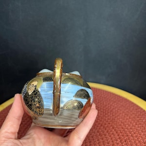 Puede incluir: Jarra de cerámica pequeña y ornamentada con asa y borde dorados. El cuerpo presenta una escena de paisaje con figuras, agua azul y detalles dorados. El diseño recuerda al arte tradicional japonés.