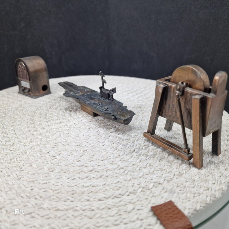 May include: A collection of miniature bronze-colored metal objects. Includes a radio, an aircraft carrier, and a sharpening wheel. The aircraft carrier has intricate details. The objects are arranged on a textured white surface.