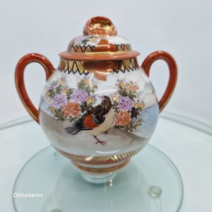 Pode incluir: Um pequeno bule de porcelana branca com tampa e alças vermelhas. O bule é decorado com um padrão floral e um pássaro em voo. O bule está sobre uma superfície de vidro.