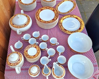 Könnte beinhalten: Ein komplettes weißes Porzellan-Geschirrset mit rotem und goldenem Zierrand. Das Set umfasst Teller, Schalen, Tassen, Untertassen, eine Teekanne, eine Servierschüssel und Servierkannen. Die Teile sind auf einer rot-weiß gemusterten Tischdecke angeordnet.