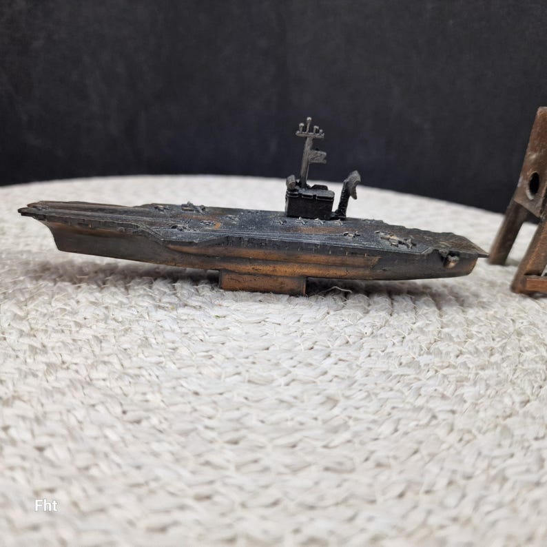 May include: A detailed miniature model of a warship, featuring a dark, weathered finish with hints of brown and rust. The model includes a detailed superstructure and a flat deck, suggesting a vintage or antique piece. The model is placed on a textured, off-white surface.