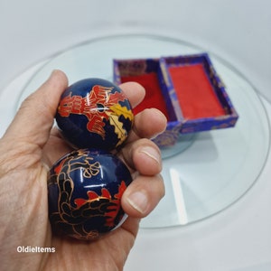 Op de afbeelding: Twee blauwe metalen ballen met ingewikkelde rode, gouden en zwarte ontwerpen. De ballen worden in een hand gehouden en worden gebruikt voor ontspanning en stressverlichting.