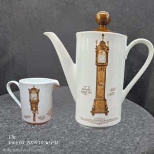 May include: A white ceramic coffee pot with a brown spherical lid and a matching creamer, both featuring a brown illustration of a tall grandfather clock and Dutch text. The coffee pot has a long spout and a handle, while the creamer has a small handle.