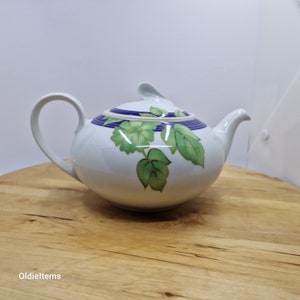 Op de afbeelding: Witte theepot met een blauwe en gouden rand en een groen bladerenontwerp.