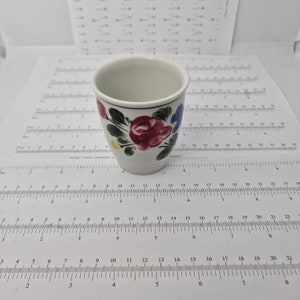 Könnte beinhalten: Eine kleine weiße Keramiktasse mit Blumenmuster. Die Tasse hat einen blauen Rand und zeigt rote, rosa und blaue Blumen mit grünen Blättern.