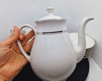 May include: White porcelain teapot with a silver trim and a lid. The teapot has a rounded body and a spout on the side. The teapot is sitting on a black surface.