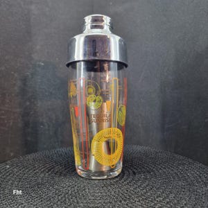 May include: A clear glass cocktail shaker with a stainless steel top and base. The glass is decorated with yellow, orange, and red graphics, including the words "Tequila Sunrise".
