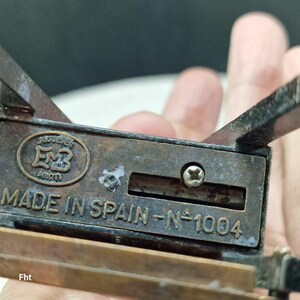 May include: Close-up of a vintage metal pencil sharpener. The sharpener is rectangular with a bronze finish and shows signs of age. The text "MADE IN SPAIN - N&deg;1004" is visible. The sharpener has the embossed letters "EMB" and the word "JUGUETES".