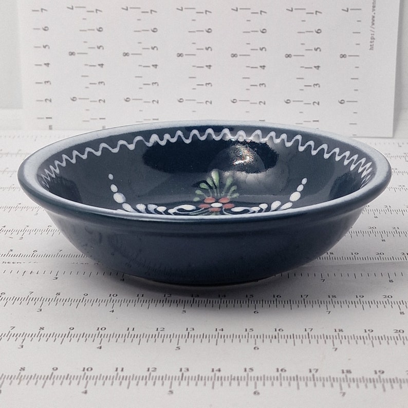 K&ouml;nnte beinhalten: Eine kleine, dunkelblaue Keramiksch&uuml;ssel mit wei&szlig;em Rand und wei&szlig;em Blumenmuster. Die Sch&uuml;ssel hat einen gewellten Rand.